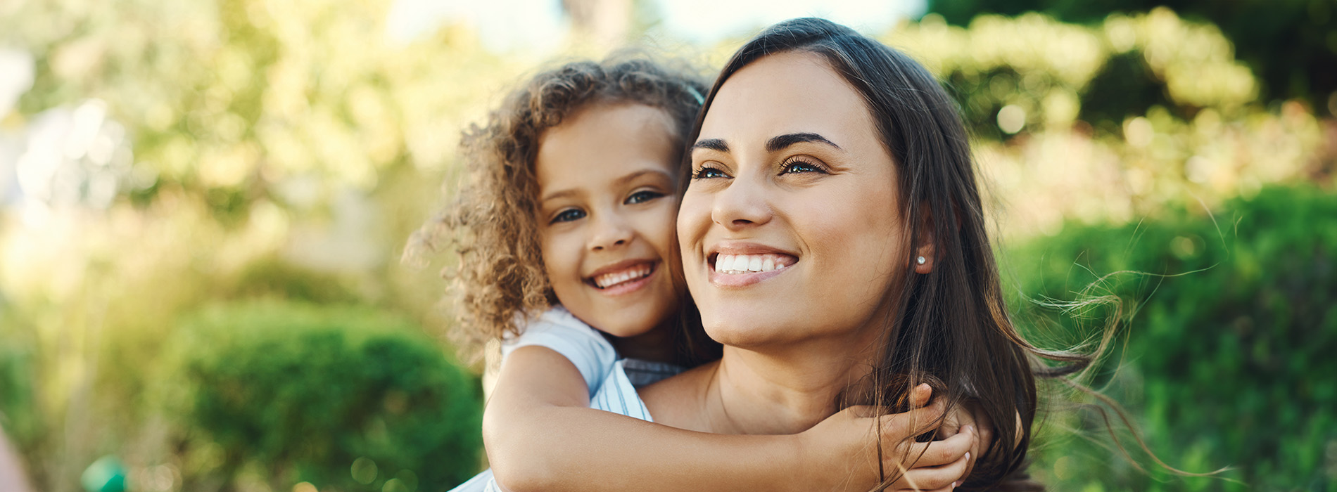 The image shows a woman holding a young child, both smiling, with a clear sky background and a partial view of another person s face.