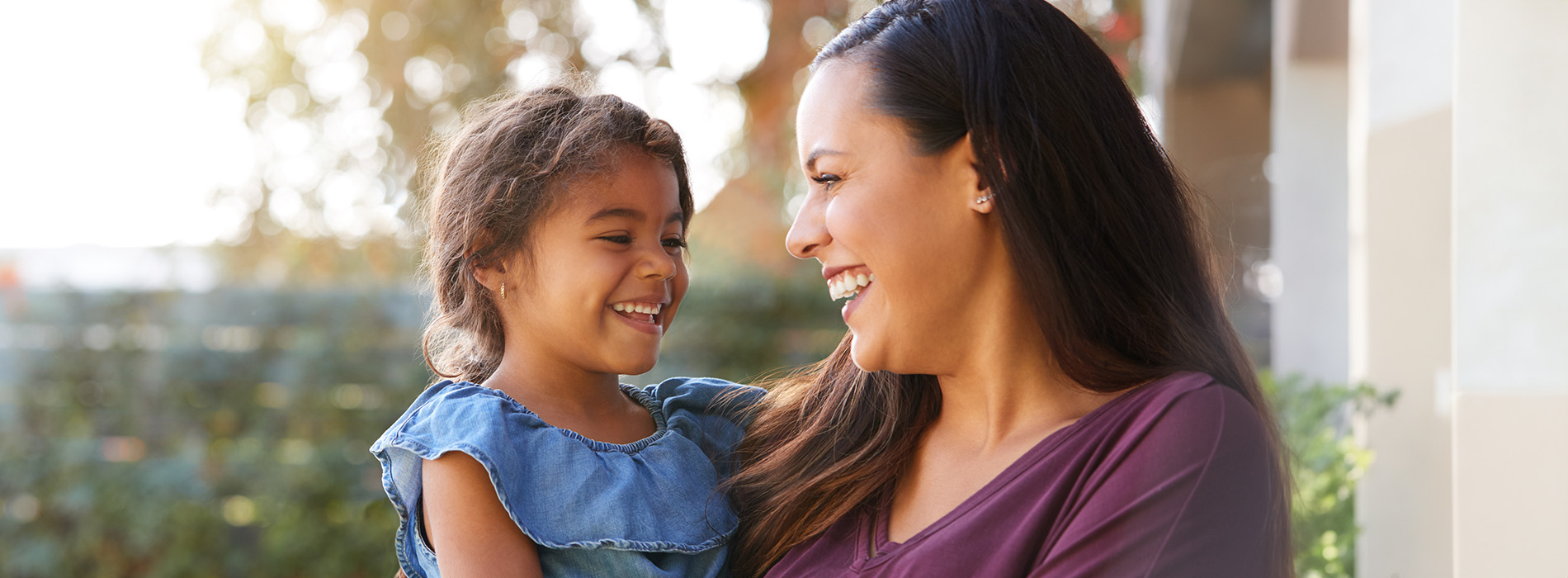 The image shows a woman and a child outdoors during daylight, with the woman holding the child and both smiling at the camera.