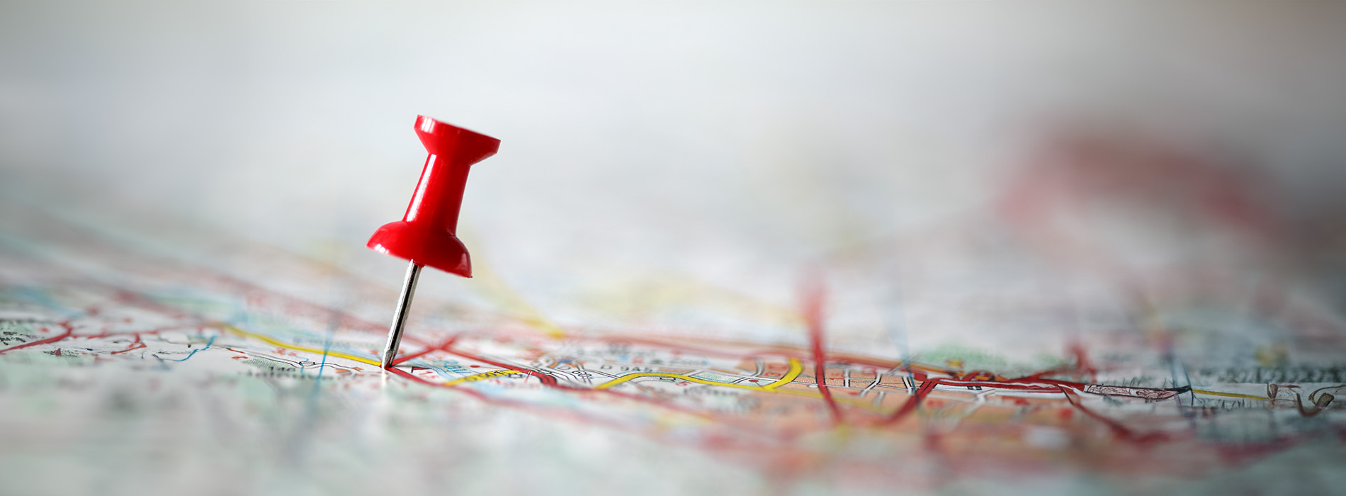 The image shows a close-up view of a map with a red pin sticking out, placed on a surface with blurred text and other objects in the background.
