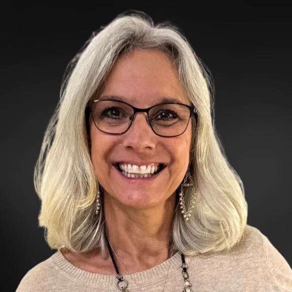 The image shows a smiling woman with gray hair, wearing glasses, a black shirt, and a necklace, standing against a dark background.