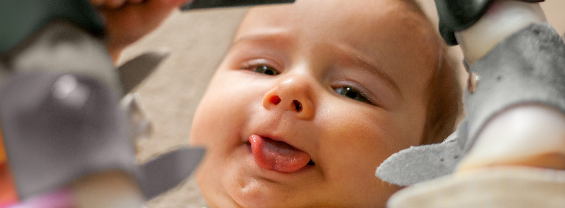 A baby with a playful expression looking at the camera while being held by an adult s hand near a small toy figure, all set against a blurred background.
