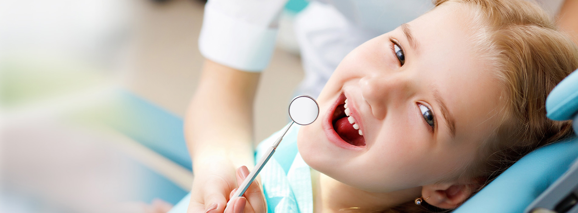 The image features a young child with blonde hair, smiling and looking at the camera, while holding a toothbrush with both hands.