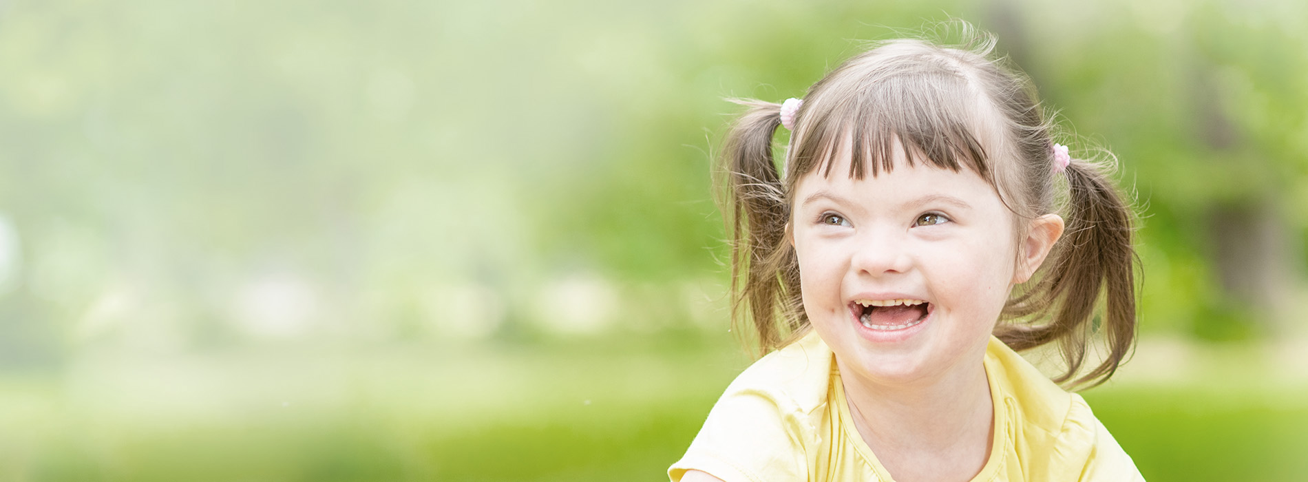 A young girl with pigtails is smiling at the camera, likely outdoors due to the natural light and greenery in the background.