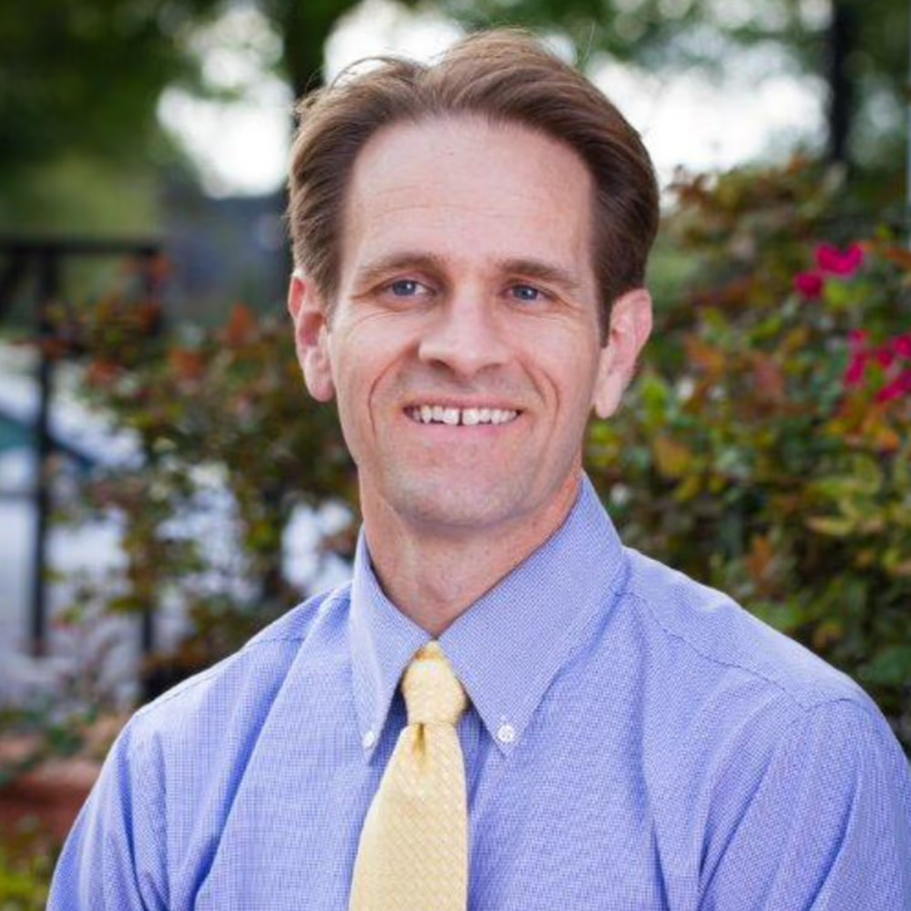 The image shows a man with short brown hair, wearing a blue shirt, yellow tie, and a light blue lanyard, posing for a portrait with a smile against a blurred background that includes trees and a fence.