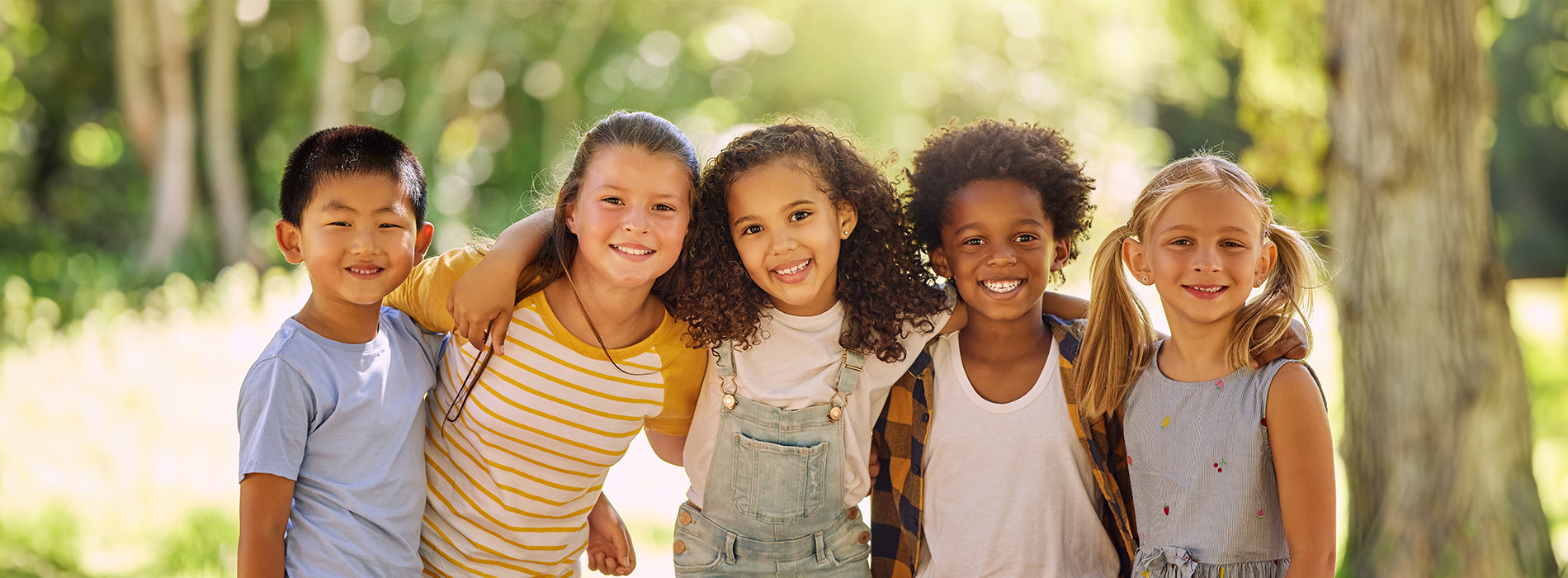 The image shows a group of children posing together outdoors with a natural background, smiling and looking towards the camera.