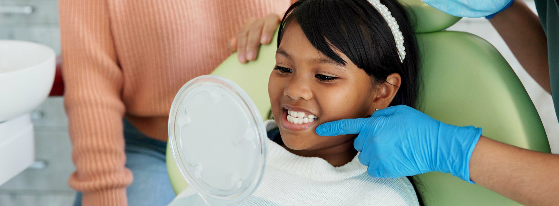 The image shows a young child sitting in a dental chair with their eyes closed, receiving dental care from a professional in a dental office setting, with a smiling face and a relaxed posture.