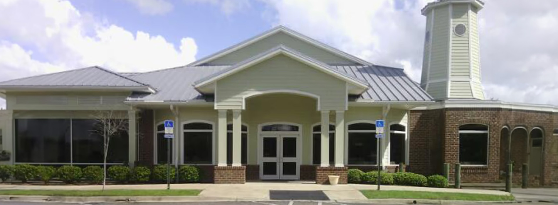 The image shows a building with a prominent entrance, possibly an office or community center, featuring a covered porch area with columns and a clock tower in the background under a clear sky.