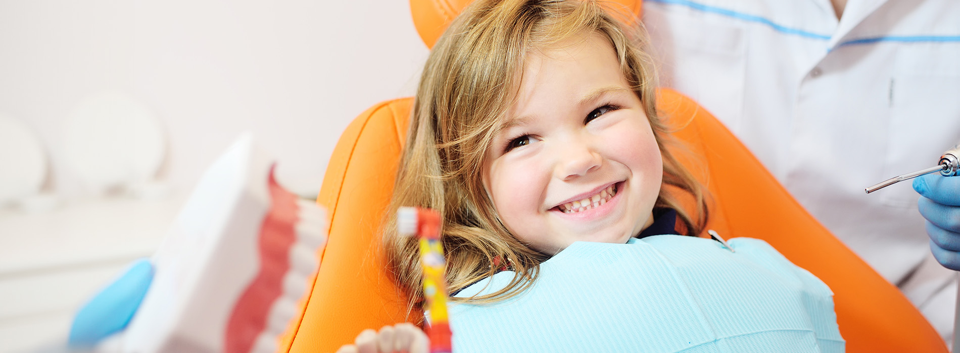 A young child sitting in an orange chair, smiling at the camera, with medical professionals in the background.