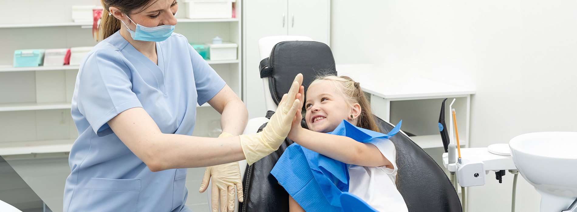 A dental hygienist is assisting a child with dental care, both wearing face masks.