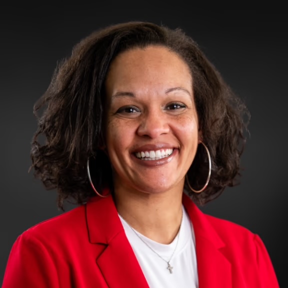 The image shows a woman with short hair wearing a red blazer and smiling at the camera.
