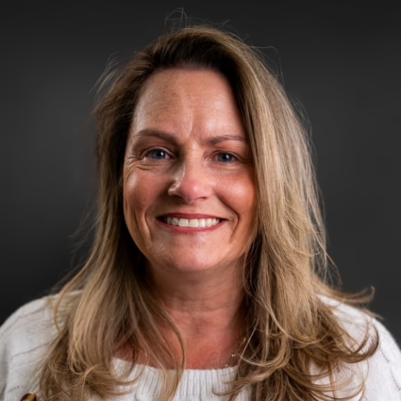 The image shows a woman with blonde hair smiling at the camera, wearing a white top and sitting against a dark background.