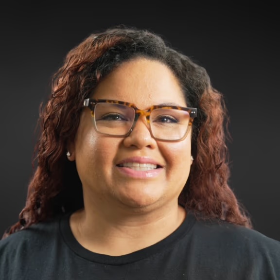 The image displays a smiling woman with glasses, wearing a dark top, against a black background.