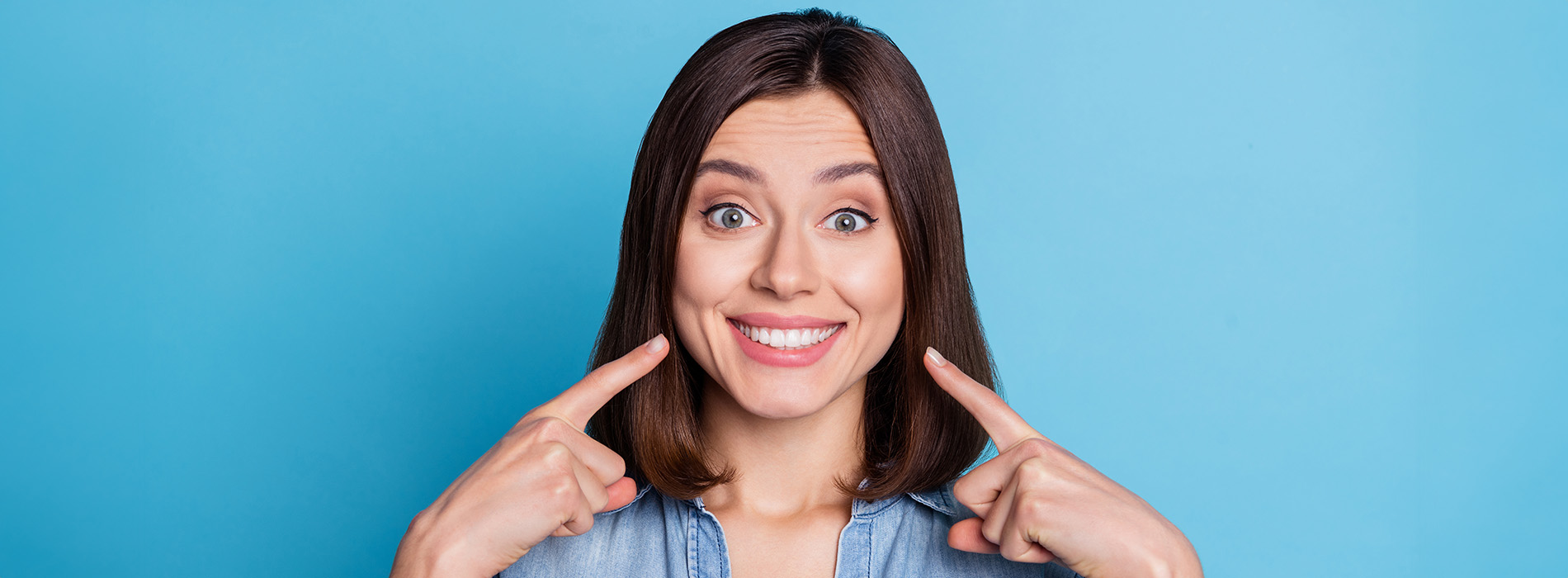 The image shows a woman with dark hair smiling at the camera, making a playful gesture with her hand near her face, against a blue background.
