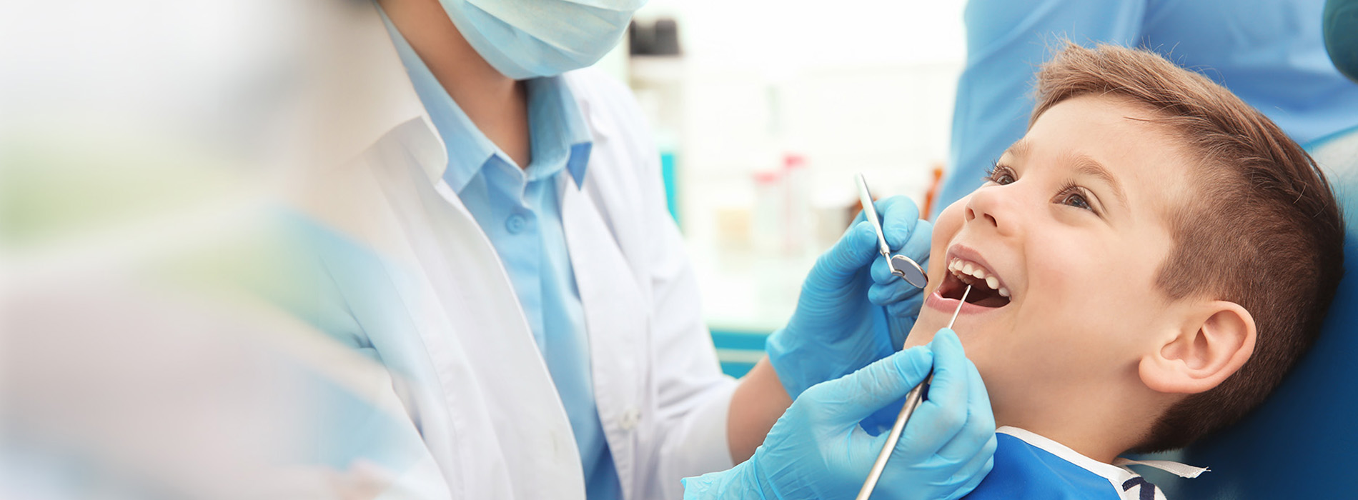 A young child receiving dental care from a dentist, with a dental hygienist assisting, all wearing protective gear.