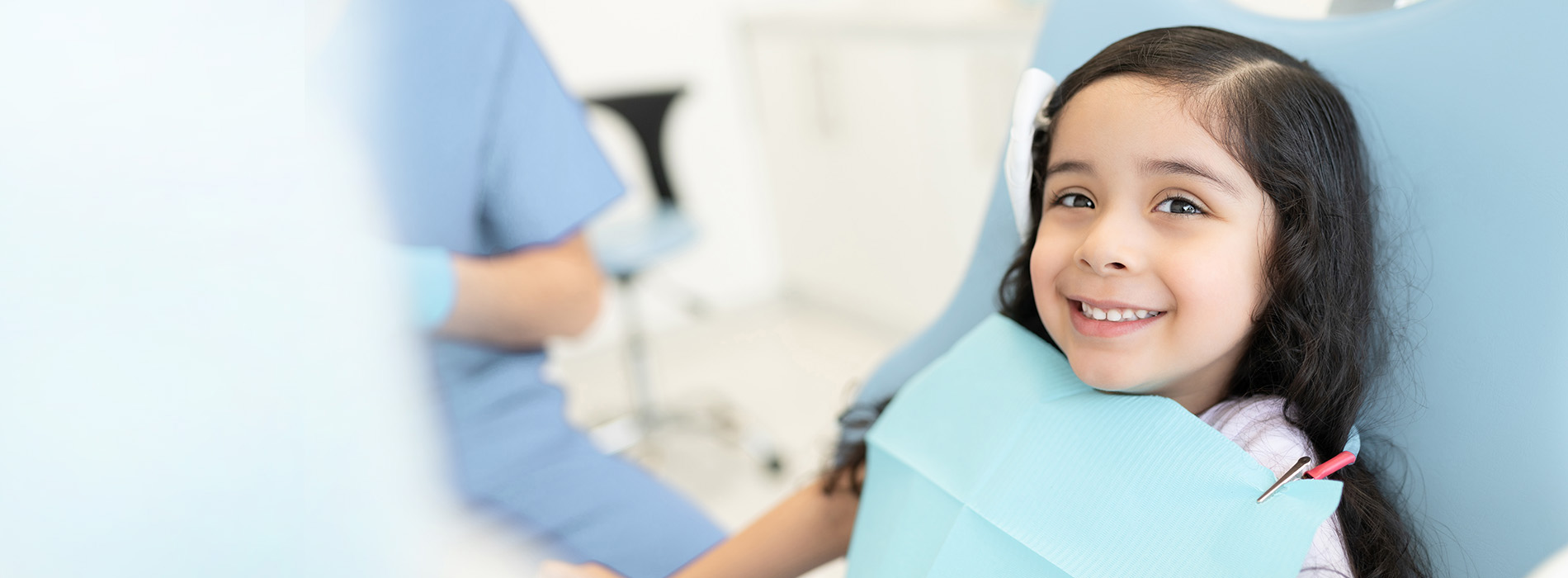 A young girl sitting in a dental chair with a smile on her face, wearing a blue bib around her neck, while a dentist works at a dental station.