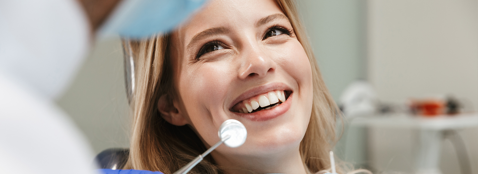 A woman with blonde hair is sitting in a dental chair, smiling, while a dentist works on her teeth.