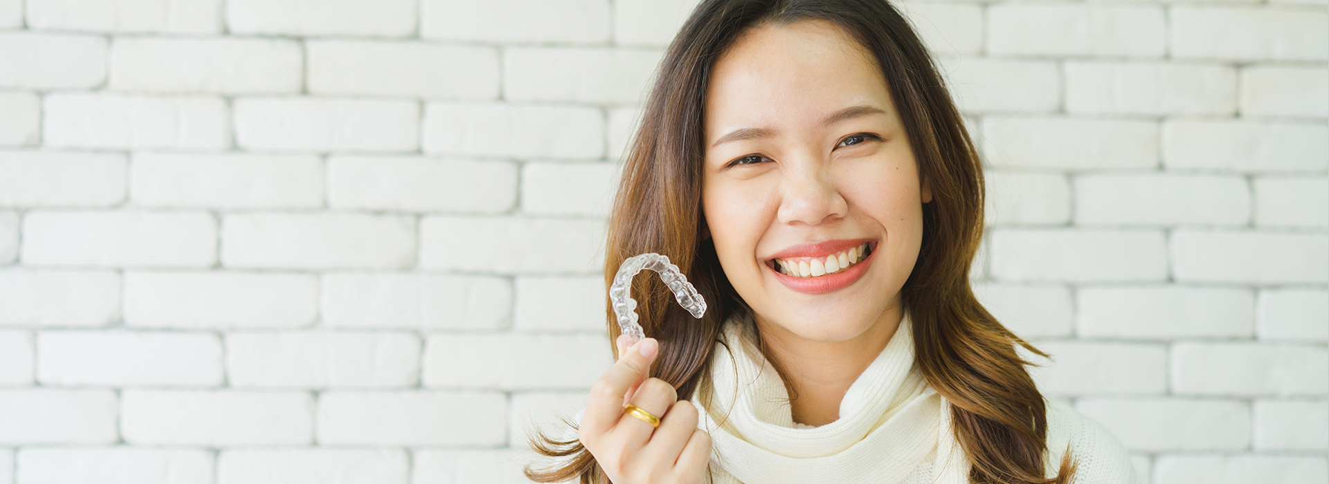 A woman holding a toothbrush with a smile while standing against a brick wall.