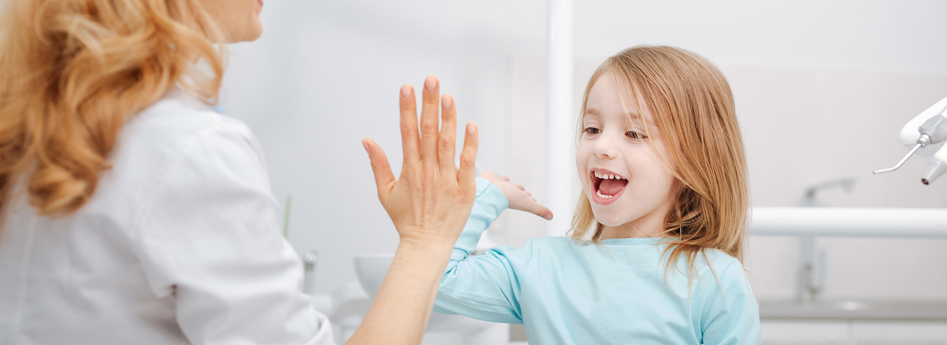 A young girl playfully waving at her reflection in a mirror with an adult behind her.
