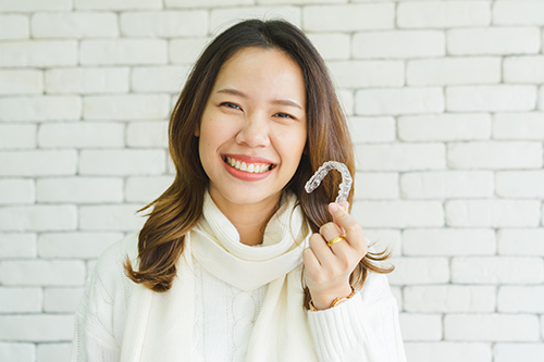 A smiling woman holding up a white peace sign with her right hand against a brick wall background.